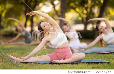 Girls practicing hatha yoga in summer garden, stretching in an asymmetrical seated asana Parivritta Janu Sirsasana with a forward bend and an outstretched arm Girls practicing hatha yoga in summer garden, stretching in an asymmetrical seated asana Parivritta Janu Sirsasana with a forward bend and an outstretched arm 122158736