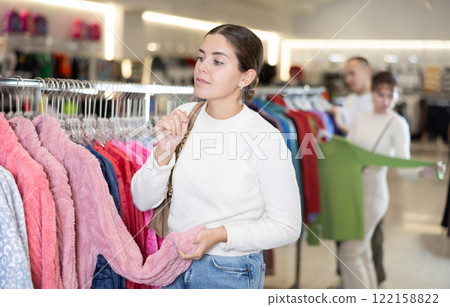 Girl stopped near leisure wear showcase, holds eiderdown bathrobe hanger in hands, examines product 122158822