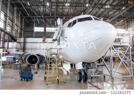 White passenger airplane in the aviation hangar. Airliner under maintenance. Checking mechanical systems for flight operations 122161375