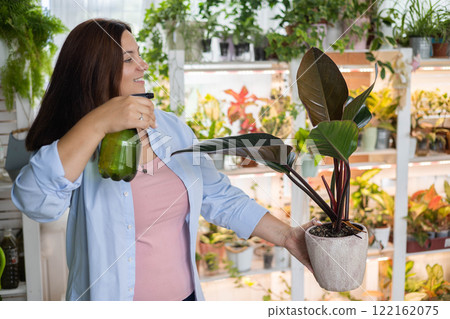 Smiling woman florist gardener sprinkling watering philodendron at indoor home garden closeup Smiling woman florist gardener sprinkling watering philodendron at indoor home garden closeup 122162075