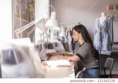 A plus size woman fashion designer uses an overlock sewing machine in her studio.  122162126
