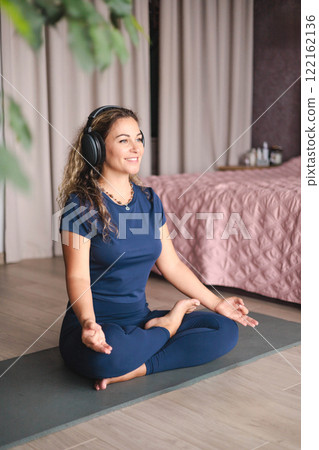 Smiling yoga woman meditation in lotus position listening music use headphones at home Smiling yoga woman meditation in lotus position listening music use headphones at home 122162136