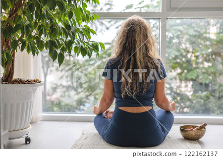 Woman yoga meditation sitting in lotus position near panorama window with houseplant at home Woman yoga meditation sitting in lotus position near panorama window with houseplant at home 122162137