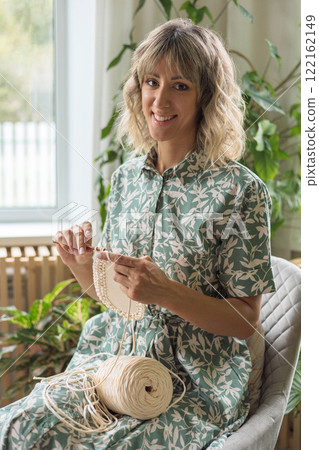 Young beautiful European girl crochets a storage basket from beige yarn. Young beautiful European girl crochets a storage basket from beige yarn. 122162149