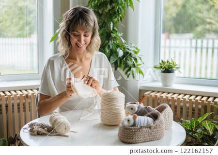 Young beautiful smiling woman sits at the table and crochets from beige yarn. Young beautiful smiling woman sits at the table and crochets from beige yarn. 122162157