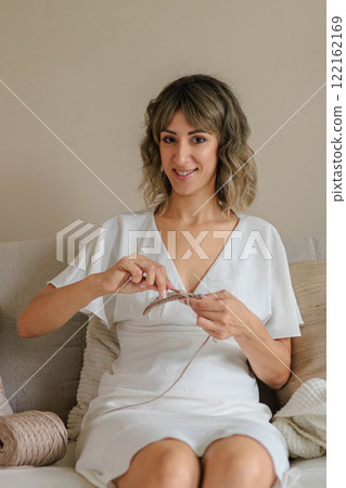 Young beautiful woman sits on the sofa and crochets from beige yarn. The girl enjoys the hobby. Young beautiful woman sits on the sofa and crochets from beige yarn. The girl enjoys the hobby. 122162169