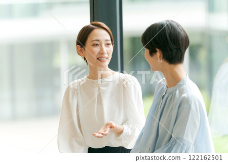 Two women talking by a window in an office 122162501