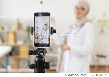 Middle-eastern adult female doctor recording video lesson on medical procedure. Demonstrates how to use a dropper correctly via smartphone. Educational content for healthcare professionals. Middle-eastern adult female doctor recording video lesson on medical procedure. Demonstrates how to use a dropper correctly via smartphone. Educational content for healthcare professionals. 122163734