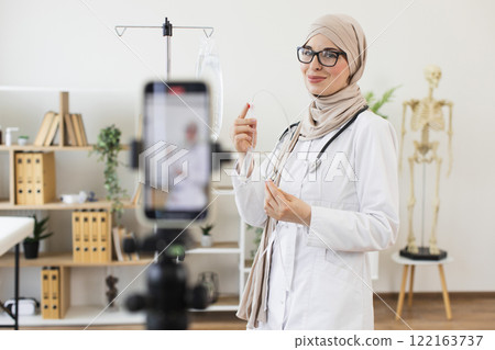 Middle-aged Muslim woman doctor explains medical dropper procedure on video using smartphone in office setting. Middle-aged Muslim woman doctor explains medical dropper procedure on video using smartphone in office setting. 122163737