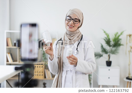Muslim female doctor recording video for blog about medicine treatment. Woman holding bottles of medicine while explaining healthcare tips to online followers. Muslim female doctor recording video for blog about medicine treatment. Woman holding bottles of medicine while explaining healthcare tips to online followers. 122163792