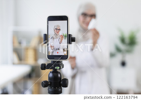 Muslim woman in hijab demonstrating medicine bottles while recording video. Young adult female creating educational content on healthcare. Professional setting suggests engaging online audience. Muslim woman in hijab demonstrating medicine bottles while recording video. Young adult female creating educational content on healthcare. Professional setting suggests engaging online audience. 122163794