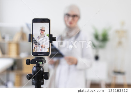 Muslim woman doctor, in her 30s, records video tutorial discussing x-ray treatment. She uses smartphone while wearing hijab and doctor's coat in home office. 122163812