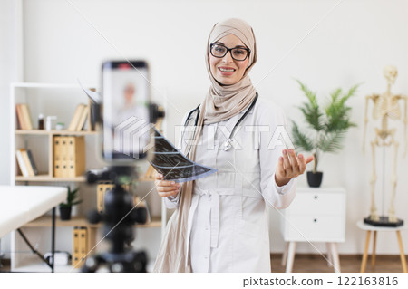 Muslim woman doctor recording video tutorial on x-ray treatment using smartphone. Middle Eastern female healthcare professional in medical office. Sharing health knowledge online. Muslim woman doctor recording video tutorial on x-ray treatment using smartphone. Middle Eastern female healthcare professional in medical office. Sharing health knowledge online. 122163816