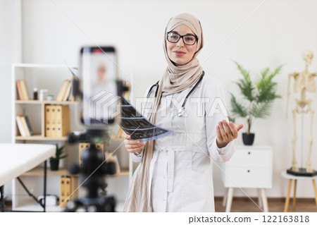 Muslim woman doctor recording educational video with x-ray, highlighting medical treatment. Female professional showcases modern healthcare approach, creating online content for patient education. Muslim woman doctor recording educational video with x-ray, highlighting medical treatment. Female professional showcases modern healthcare approach, creating online content for patient education. 122163818