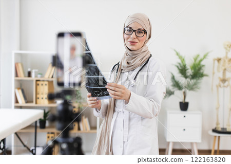 Muslim woman doctor recording educational video with x-ray, highlighting medical treatment. Female professional showcases modern healthcare approach, creating online content for patient education. Muslim woman doctor recording educational video with x-ray, highlighting medical treatment. Female professional showcases modern healthcare approach, creating online content for patient education. 122163820