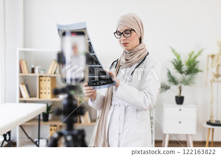 Muslim woman doctor in hijab recording video lesson on x-ray analysis using smartphone. Middle-Eastern, adult, professional wearing lab coat, sharing medical knowledge. 122163822