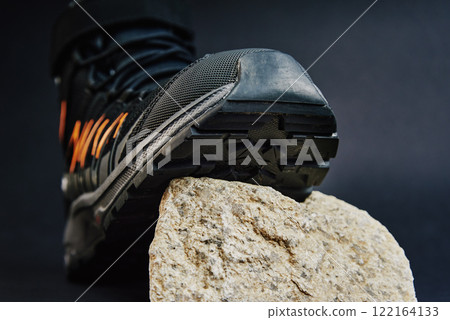 Close-up of waterproof black hiking shoe with orange details on small rock. Rugged trekking boot on dark background. Footwear for exploration and outdoor activities 122164133