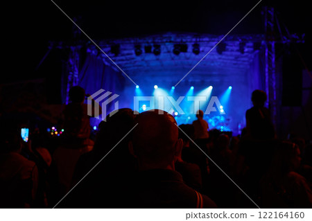 Silhouettes of crowd at concert in front of stage with bright spotlights. People enjoy live music at the band's performance. Concert hall with musicians on stage and fans during music festival Silhouettes of crowd at concert in front of stage with bright spotlights. People enjoy live music at the band's performance. Concert hall with musicians on stage and fans during music festival 122164160