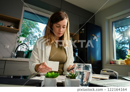 Stressed woman reviewing bills at home. Female organizing receipts, sitting at kitchen table with laptop and paper documents. Concept of budgeting and managing personal finances Stressed woman reviewing bills at home. Female organizing receipts, sitting at kitchen table with laptop and paper documents. Concept of budgeting and managing personal finances 122164164