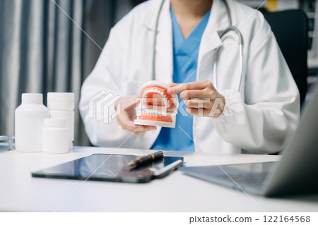 Dentist sitting at table with jaw samples tooth model and working with tablet and laptop in dental clinic. 122164568