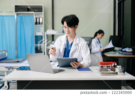 Confident young male doctor in white medical uniform sit at desk working on computer Confident young male doctor in white medical uniform sit at desk working on computer 122165000