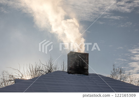 A house chimney emitting smoke on a winter day, with the roof covered in snow A house chimney emitting smoke on a winter day, with the roof covered in snow 122165309