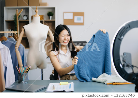 Asian lady blogger showing clothes in front of the camera to recording vlog video live streaming at her shop.  122165740