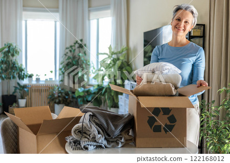 Gray haired mature smiling woman pack clothes for recycling to cardboard box at home Gray haired mature smiling woman pack clothes for recycling to cardboard box at home 122168012
