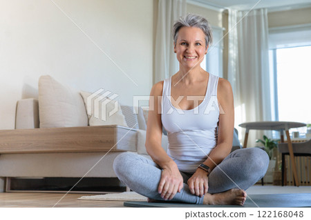 An elderly smiling yogini woman with gray hair sits in the lotus position and meditates at home. An elderly smiling yogini woman with gray hair sits in the lotus position and meditates at home. 122168048