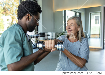 Man doctor providing physiotherapy and rehabilitation with dumbbells to adult woman Man doctor providing physiotherapy and rehabilitation with dumbbells to adult woman 122168261