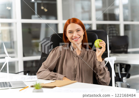 Pretty smiling ginger young woman with apple in hands sitting in the office 122168484