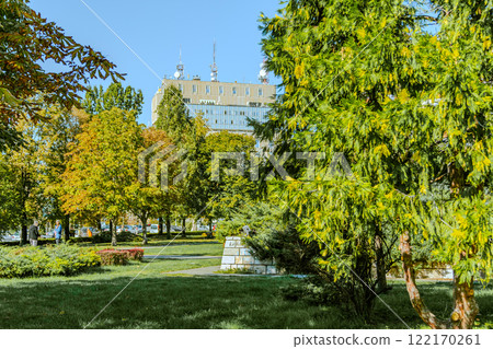 Tranquil wooden bench along the path of a city park. Grove of mature deciduous trees with green lawn make it a magical place to rest. Tranquil wooden bench along the path of a city park. Grove of mature deciduous trees with green lawn make it a magical place to rest. 122170261