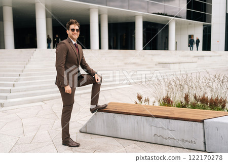 Full length portrait of happy successful businessman in brown suit and sunglasses standing in front of modern office building. Photogenic businessman in elegant formal attire posing alone outdoors. Full length portrait of happy successful businessman in brown suit and sunglasses standing in front of modern office building. Photogenic businessman in elegant formal attire posing alone outdoors. 122170278