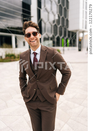 Vertical portrait of cheerful businessman wearing stylish sunglasses and suit standing posing at city, smiling looking at camera, holding hands in pockets, exuding professionalism and leadership. 122170279