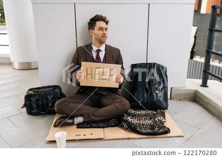 Homeless millennial businessman in suit sitting on city street holding sign HELP, illustrating hardships of poverty and unemployment. Concept of support in financial crises. 122170280