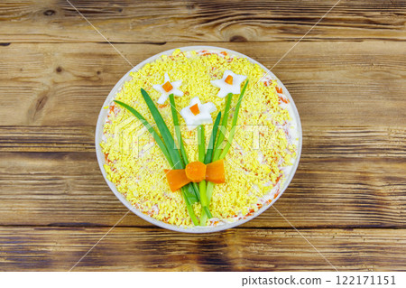 Traditional russian layered salad Mimosa with spring decoration Daffodil on wooden table. Top view. Decoration is made of egg, green onion and carrot 122171151