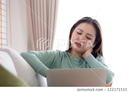 Asian woman sitting on sofa with neck pain while using laptop computer in living room at home. Asian woman sitting on sofa with neck pain while using laptop computer in living room at home. 122171533