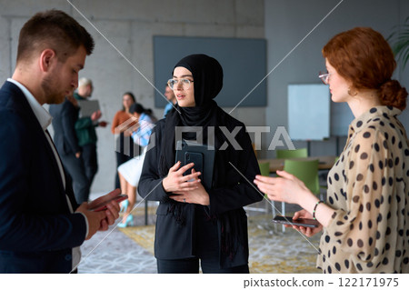 A diverse group of business professionals engages in a conversation during a work break in the office, with their colleagues visible in the background, fostering collaboration and connection A diverse group of business professionals engages in a conversation during a work break in the office, with their colleagues visible in the background, fostering collaboration and connection 122171975