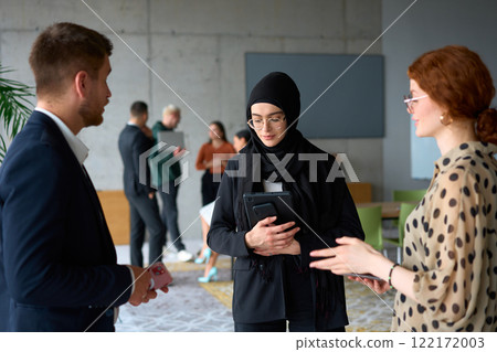 A diverse group of business professionals engages in a conversation during a work break in the office, with their colleagues visible in the background, fostering collaboration and connection 122172003