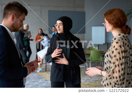 A diverse group of business professionals engages in a conversation during a work break in the office, with their colleagues visible in the background, fostering collaboration and connection 122172022