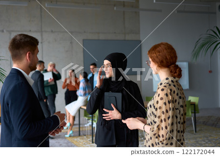A diverse group of business professionals engages in a conversation during a work break in the office, with their colleagues visible in the background, fostering collaboration and connection 122172044