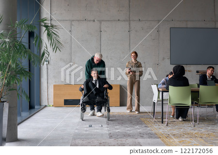 A diverse team of business colleagues, including a director in a wheelchair, engages in a collaborative discussion as they move together through a modern office space A diverse team of business colleagues, including a director in a wheelchair, engages in a collaborative discussion as they move together through a modern office space 122172056