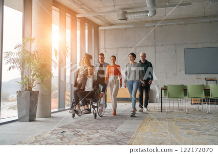 A team ofbusiness professionals moves through a spacious, modern office, assisting their leader, a businessman in a wheelchair, as they collaborate and navigate the workplace together. 122172070