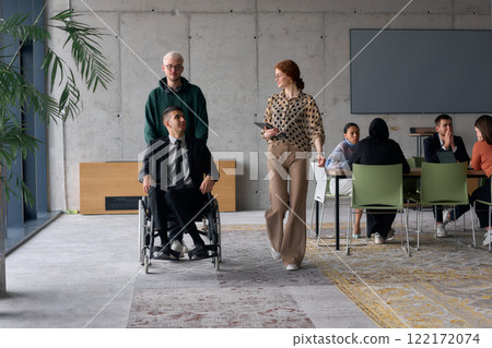 A diverse team of business colleagues, including a director in a wheelchair, engages in a collaborative discussion as they move together through a modern office space 122172074