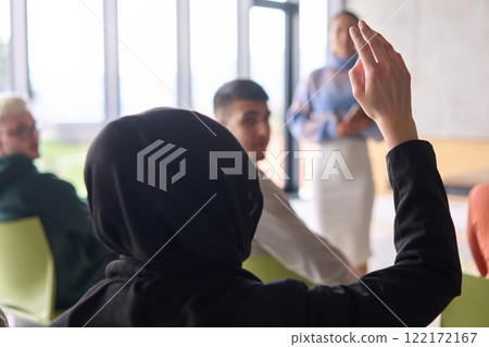 A Muslim businesswoman, dressed in professional attire and wearing a hijab, raises her hand during a business meeting to ask a question to the educator leading the session 122172167