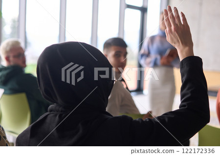 A Muslim businesswoman, dressed in professional attire and wearing a hijab, raises her hand during a business meeting to ask a question to the educator leading the session 122172336