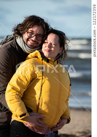 A couple stands embraced on a sandy beach on a chilly autumn day, the serene ocean stretching behind them as they share a quiet moment of love and warmth amidst the cool breeze and peaceful A couple stands embraced on a sandy beach on a chilly autumn day, the serene ocean stretching behind them as they share a quiet moment of love and warmth amidst the cool breeze and peaceful 122172359