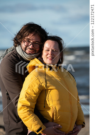 A couple stands embraced on a sandy beach on a chilly autumn day, the serene ocean stretching behind them as they share a quiet moment of love and warmth amidst the cool breeze and peaceful 122172471