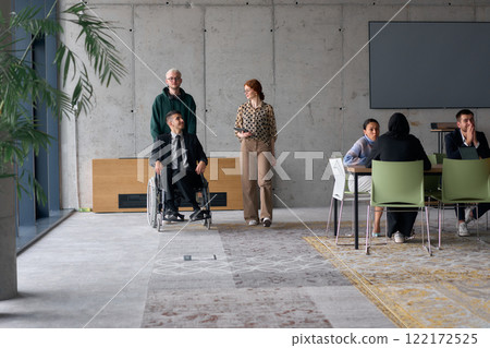 A diverse team of business colleagues, including a director in a wheelchair, engages in a collaborative discussion as they move together through a modern office space A diverse team of business colleagues, including a director in a wheelchair, engages in a collaborative discussion as they move together through a modern office space 122172525