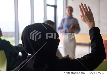 A Muslim businesswoman, dressed in professional attire and wearing a hijab, raises her hand during a business meeting to ask a question to the educator leading the session 122172532